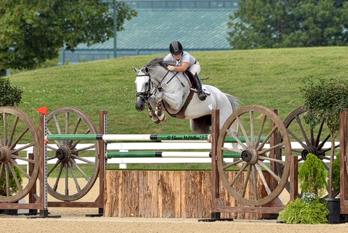 Grey mare and rider jump a large fence with a natural base, green and white striped poles, and giant wagon wheel standards. 
