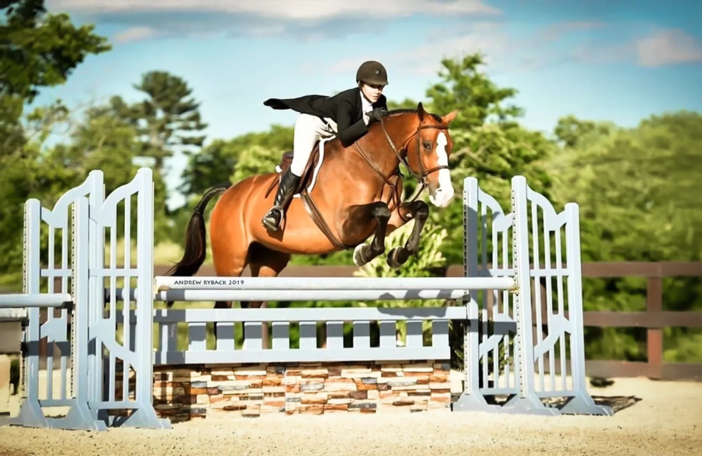 Bay horse with big blaze and rider in formal show attire jump a grey fence with a brick colored box at the base.