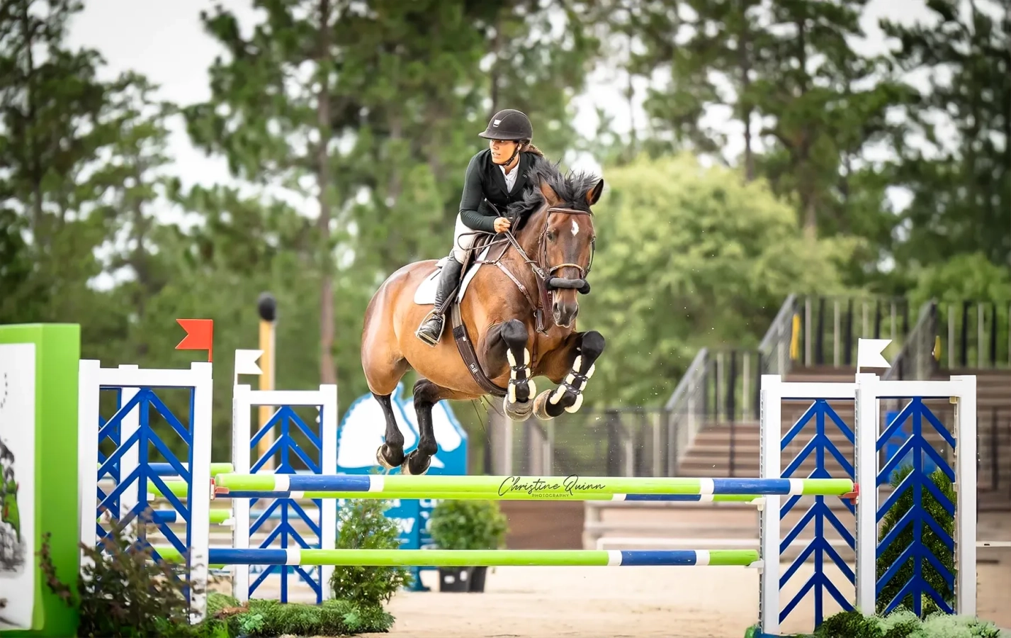 Bay horse wearing fleece-lined open front boots jumps a large fence will rider looks ahead to their next obstacle.