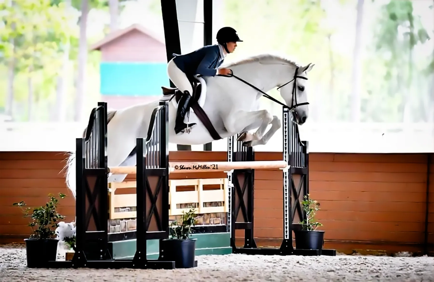 Side view of a grey horse and rider in show attire jumping an oxer with black standards in a covered arena at a horse show.